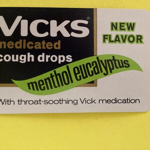 May include: A rectangular package for Vicks medicated cough drops. The package has a black background with white text that reads "Vicks medicated cough drops." A green leaf shape with "menthol eucalyptus" is in the center. The right side is white with "NEW FLAVOR" in green.