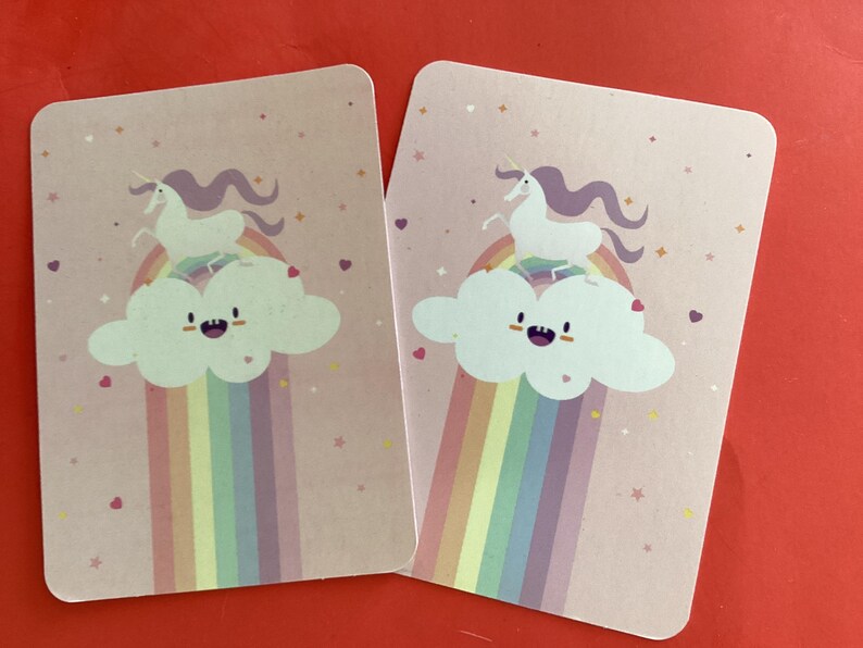 May include: Two rectangular cards with rounded corners. Each card features a cartoon unicorn atop a rainbow and cloud on a pink background. The unicorn is white with a purple mane. The rainbow has multiple colors.