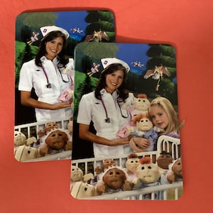 May include: Two rectangular Cabbage Patch Kids themed images. Each features a woman in a nurse's uniform holding a doll, with a child and more dolls in the background. The background shows a forest scene.