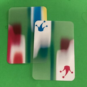 May include: Two playing cards with a translucent appearance. One card features a blue crown, the other a red jester hat. Both cards have a blurred background of green, white, red, blue, and yellow.