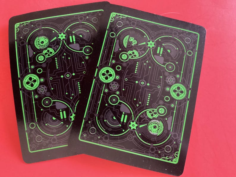 Puede incluir: Dos naipes con fondo negro y dise&ntilde;o de placa de circuito verde. El dise&ntilde;o incluye engranajes, c&iacute;rculos y l&iacute;neas. Las cartas est&aacute;n sobre una superficie roja.