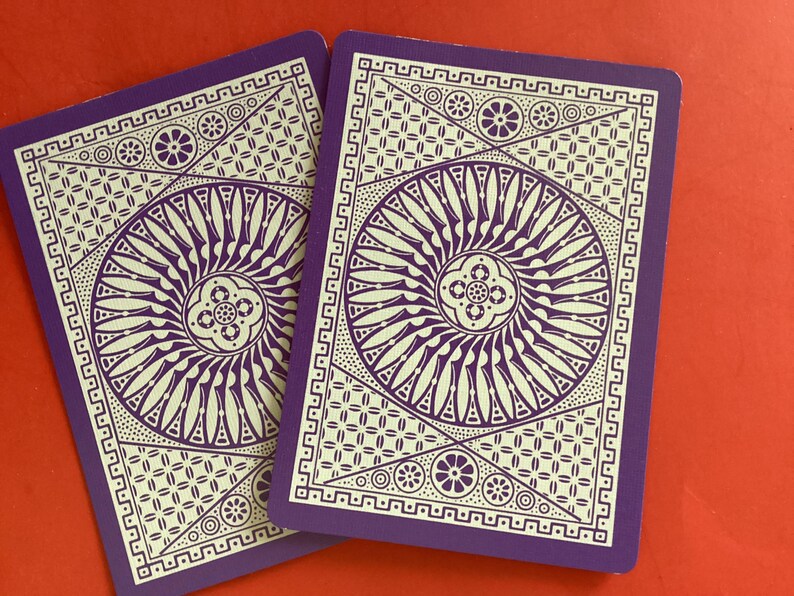 K&ouml;nnte beinhalten: Zwei Spielkarten mit einem lila Hintergrund und einem wei&szlig;en, komplizierten Design. Das Design zeigt ein zentrales kreisf&ouml;rmiges Muster mit radialen Linien, umgeben von geometrischen Formen und floralen Elementen. Die Karten sind rechteckig mit abgerundeten Ecken.