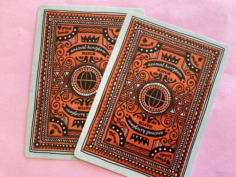 May include: Two playing cards with an orange and black design on a light blue background. The cards feature the words "animal kingdom" and "HATCH CA". The design includes a globe, crown, and decorative swirls.