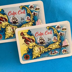 May include: Two rectangular Cape Cod souvenir coasters. Each coaster features a map of Cape Cod with illustrations of a lobster, a sailboat, and other landmarks. The words "Cape Cod" are written in red script.