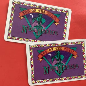 以下が含まれることがあります： 「END OF THE TRAIL NEW MEXICO」の文字が入った長方形のカードが2枚。カードは紫色の背景に、カウボーイが馬に乗ったデザイン。ボーダーは白で、黄色、赤、緑の三角形が描かれています。