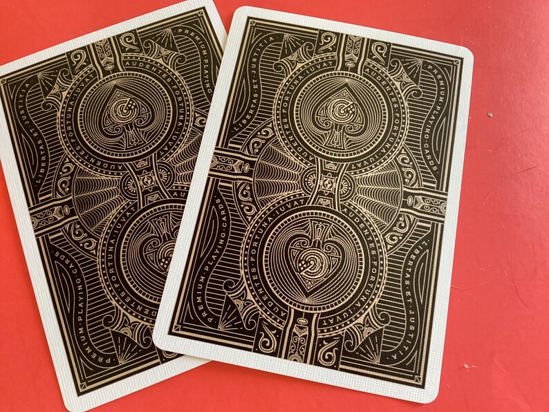 Puede incluir: Dos naipes con dise&ntilde;os ornamentados en negro y dorado, con un s&iacute;mbolo de pica en el centro. Las cartas tienen bordes blancos y est&aacute;n sobre un fondo rojo. El dise&ntilde;o incluye texto y elementos decorativos.