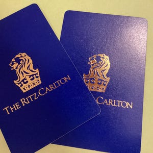 May include: Two navy blue cards with rounded corners. Each card features a gold lion crest and the words "THE RITZ-CARLTON" in gold lettering. The cards are slightly overlapping, with one partially obscuring the other.