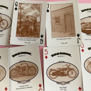 May include: A collection of Harley-Davidson playing cards featuring sepia-toned images of vintage motorcycles. Each card displays a different model and year, with text describing the motorcycle's history. The cards are arranged on a pink surface.