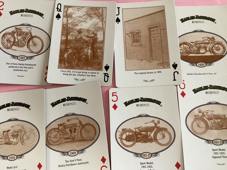 May include: A collection of Harley-Davidson playing cards featuring sepia-toned images of vintage motorcycles. Each card displays a different model and year, with text describing the motorcycle's history. The cards are arranged on a pink surface.