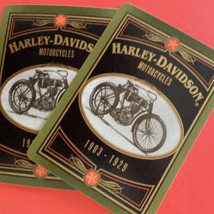 May include: Two playing cards featuring a vintage Harley-Davidson motorcycle design. The cards have a black background with the words "Harley-Davidson Motorcycles" and the years "1903-1929". The border is green with red star accents.