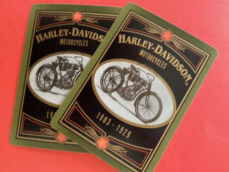May include: Two playing cards featuring a vintage Harley-Davidson motorcycle design. The cards have a black background with the words "Harley-Davidson Motorcycles" and the years "1903-1929". The border is green with red star accents.