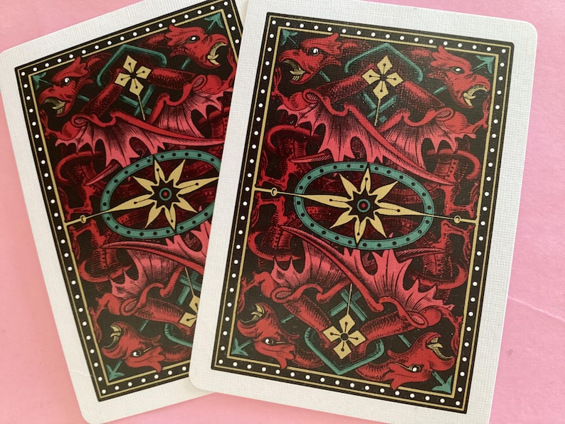 May include: Two playing cards with a detailed dragon design. The cards feature a red and black color scheme with a central compass rose design. The border is white with a dotted pattern and a gold and green frame.