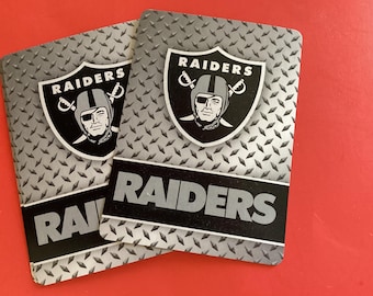 May include: Two playing cards featuring the Raiders logo. The cards have a diamond plate pattern background. The Raiders logo is a shield with a pirate wearing a helmet and the word "RAIDERS" above the logo.
