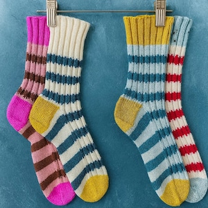 May include: Four pairs of colorful striped socks hang from a metal bar with gold clips. The socks feature various color combinations, including pink and brown, cream and teal, teal and yellow, and red and white. The socks are made of a ribbed knit material.