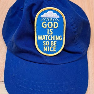 May include: A royal blue baseball cap featuring a centered oval patch with a yellow border. The patch displays a white cloud icon above the yellow text "GOD IS WATCHING SO BE NICE" set against a blue background, with a button on the crown.