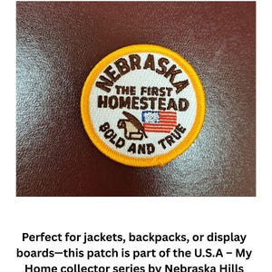 May include: A circular embroidered patch with a bright yellow border and white background. It features the text NEBRASKA, THE FIRST HOMESTEAD, and BOLD AND TRUE in brown. A brown plow icon and a small rectangular United States flag are centered.