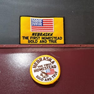 May include: A rectangular yellow embroidered patch featuring the American flag and the text NEBRASKA THE FIRST HOMESTEAD BOLD AND TRUE, alongside a circular yellow patch with the same text and a small flag graphic with a brown homestead icon.