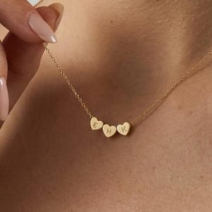 May include: A gold-colored necklace with three heart-shaped pendants. Each pendant has a single letter engraved on it. The necklace is worn around the neck.