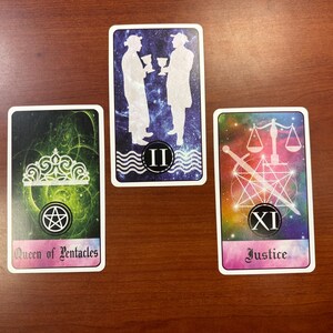 May include: Three tarot cards are displayed on a wooden surface. The cards feature illustrations of figures, symbols, and text. The cards are labeled "Queen of Pentacles", "II", and "Justice". The background of the cards have a galaxy theme.