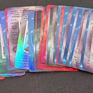 May include: A spread of tarot cards, showcasing a range of colours and mystical designs. The cards have red, blue, and green tones with iridescent highlights. Some cards feature illustrations of figures and symbols.