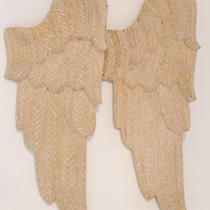 Puede incluir: Un par de alas de ángel decorativas, talladas en un material beige claro. Las alas tienen un diseño detallado de plumas y están montadas sobre un fondo blanco. Las alas son un artículo decorativo.