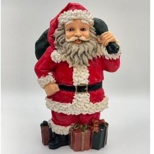May include: A Santa Claus figurine wearing a red suit with white textured trim, a black belt with a gold buckle, and a matching red hat. Santa has a long white beard and carries a dark green sack over one shoulder, standing beside several wrapped gift boxes.