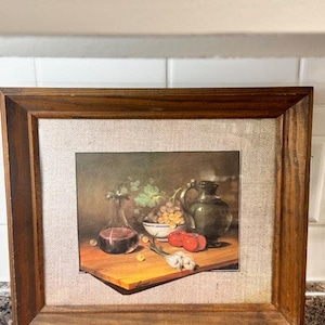 May include: A framed still life print featuring a wooden cutting board with a glass bottle of red liquid, grapes, tomatoes, and a green pitcher. The print is set against a neutral linen background within a dark wood frame.