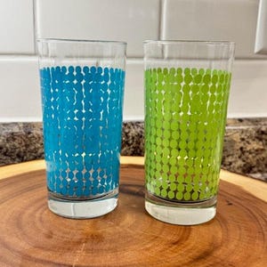 May include: Two clear drinking glasses with a dot pattern. One glass has a blue dot pattern, and the other has a green dot pattern. The glasses are cylindrical and sit on a wooden surface.