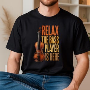 May include: Black t-shirt featuring a violin graphic and the text "RELAX THE BASS PLAYER IS HERE" in orange and yellow. The violin is brown with black strings. The shirt is made of a soft material.