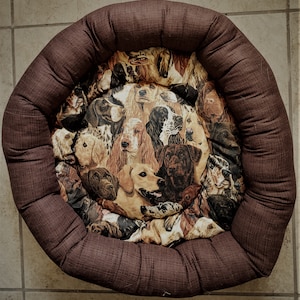 Peut inclure: Un lit pour chien marron avec un tissu à motifs représentant différentes races de chiens. Le tissu est un mélange de marron, blanc et noir.