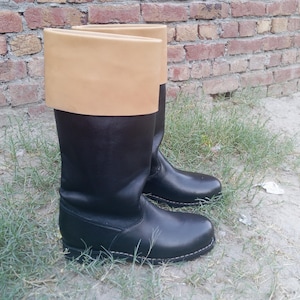 May include: A pair of knee-high boots featuring a smooth black lower section and a wide, folded-over tan cuff at the top. The boots have a rounded toe, a flat sole with visible stitching along the base, and a structured shaft that reaches the calf.