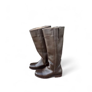 May include: A pair of brown leather riding boots. The boots are knee-high with a rounded toe and a small heel. The leather has a slightly textured appearance and a dark brown color.