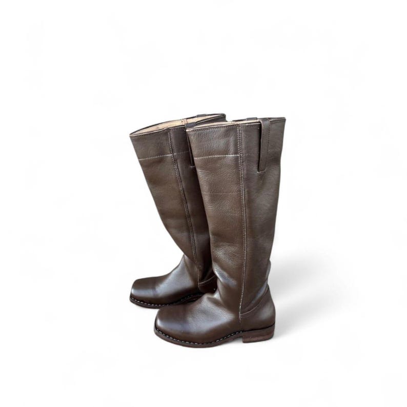 May include: A pair of brown leather riding boots. The boots are knee-high with a rounded toe and a small heel. The leather has a slightly textured appearance and a dark brown color.
