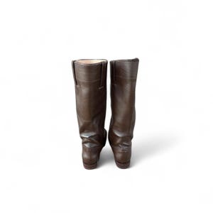 May include: A pair of brown leather riding boots. The boots are tall, with a smooth, matte finish. The boots have a classic design, with a slight heel and a rounded toe. The boots are set against a white background.