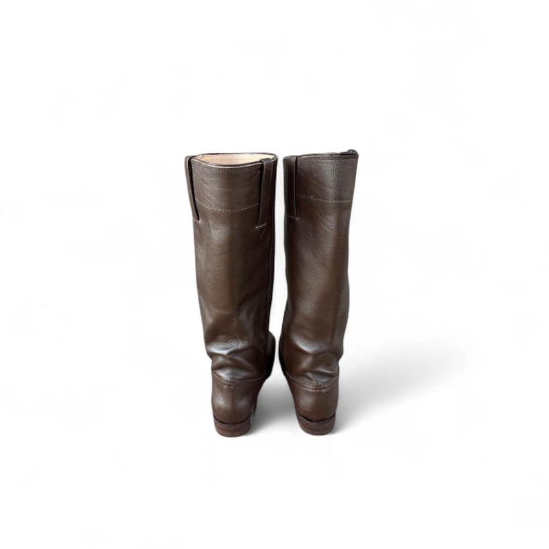 May include: A pair of brown leather riding boots. The boots are tall, with a smooth, matte finish. The boots have a classic design, with a slight heel and a rounded toe. The boots are set against a white background.
