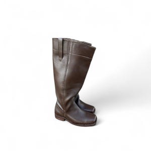 May include: A pair of brown leather knee-high boots. The boots have a square toe, a small heel, and a strap detail at the top of the boot shaft. The leather appears smooth and the boots are set against a white background.