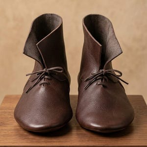 May include: A pair of dark brown leather ankle boots featuring a rounded toe, a high-cut collar with a folded-over flap design, and thin leather laces tied in a bow at the center of the vamp. The boots have a smooth, matte finish.