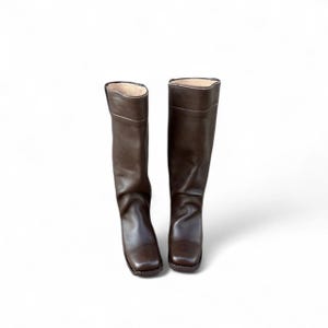 May include: A pair of brown leather riding boots. The boots are knee-high with a square toe and a slight heel. The leather has a smooth texture and a rich, dark brown color. The interior lining is a lighter tan color.