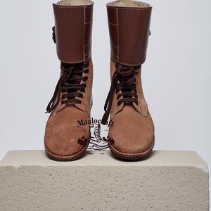 May include: Two brown suede boots with dark brown leather cuffs and laces. The boots feature a classic design with a buckle detail. The boots are displayed on a light-coloured surface. The text "Maalocraft" is visible on the boots.
