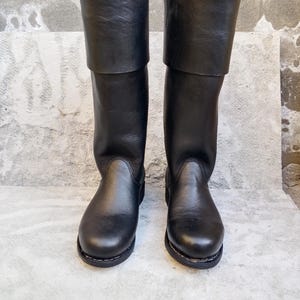May include: A pair of black leather boots with a folded-over cuff. The boots have a rounded toe and a sturdy sole. The leather appears smooth and has a slight sheen. The boots are standing upright on a light gray surface.