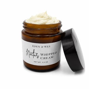 Op de afbeelding: Een klein, bruin glazen potje gevuld met witte slagroom. De pot heeft een zwarte deksel ernaast. Het etiket luidt "Nectar Whipped Cream" en "Netto gewicht 99 g".