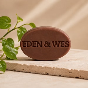 May include: An oval-shaped brown bar of soap featuring the embossed text EDEN & WES on its front surface. The soap has a smooth, solid texture and is positioned upright, displaying clear lettering centred on the face of the brown bar.