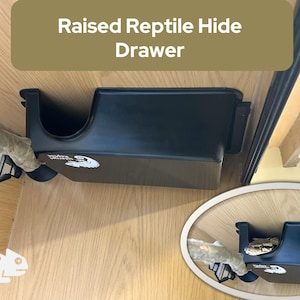 May include: A black reptile hide drawer with the text "Raised Reptile Hide Drawer". The drawer is mounted inside a wooden enclosure. A branch is placed inside the drawer. A smaller, oval-shaped image shows the drawer with a reptile inside.