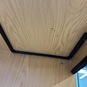 May include: A close-up view of the underside of a wooden structure. The light-colored wood grain is visible. A black metal bracket is attached to the wood with screws. The bracket forms a right angle.
