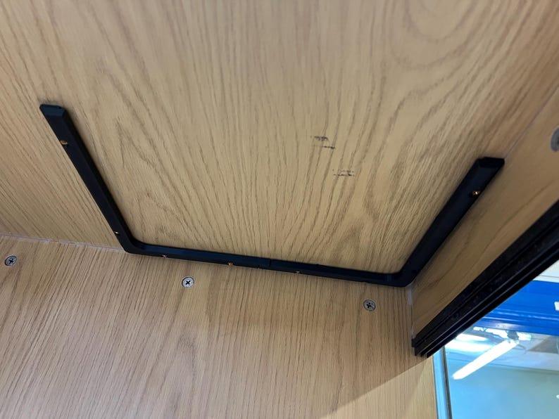 May include: A close-up view of the underside of a wooden structure. The light-colored wood grain is visible. A black metal bracket is attached to the wood with screws. The bracket forms a right angle.