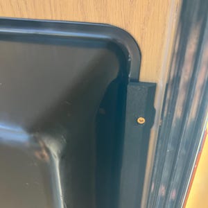 May include: Close-up of a black metal tray with rounded corners. A black metal bracket is attached to the tray and a wooden surface. A small gold-colored screw secures the bracket to the wooden surface.