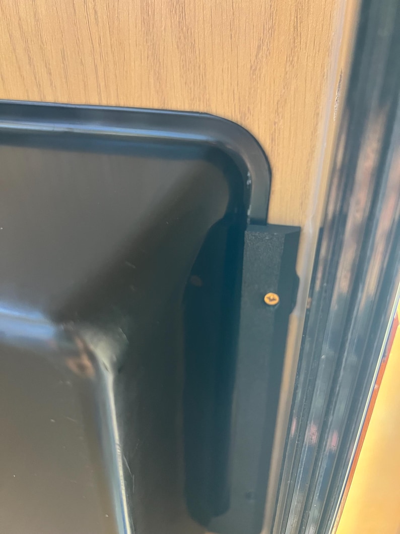 May include: Close-up of a black metal tray with rounded corners. A black metal bracket is attached to the tray and a wooden surface. A small gold-colored screw secures the bracket to the wooden surface.