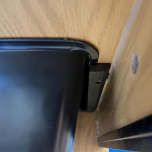May include: A black, rectangular container with rounded corners is mounted under a wooden surface. A black bracket is attached to the container and the underside of the wood. The wood has a light brown color.