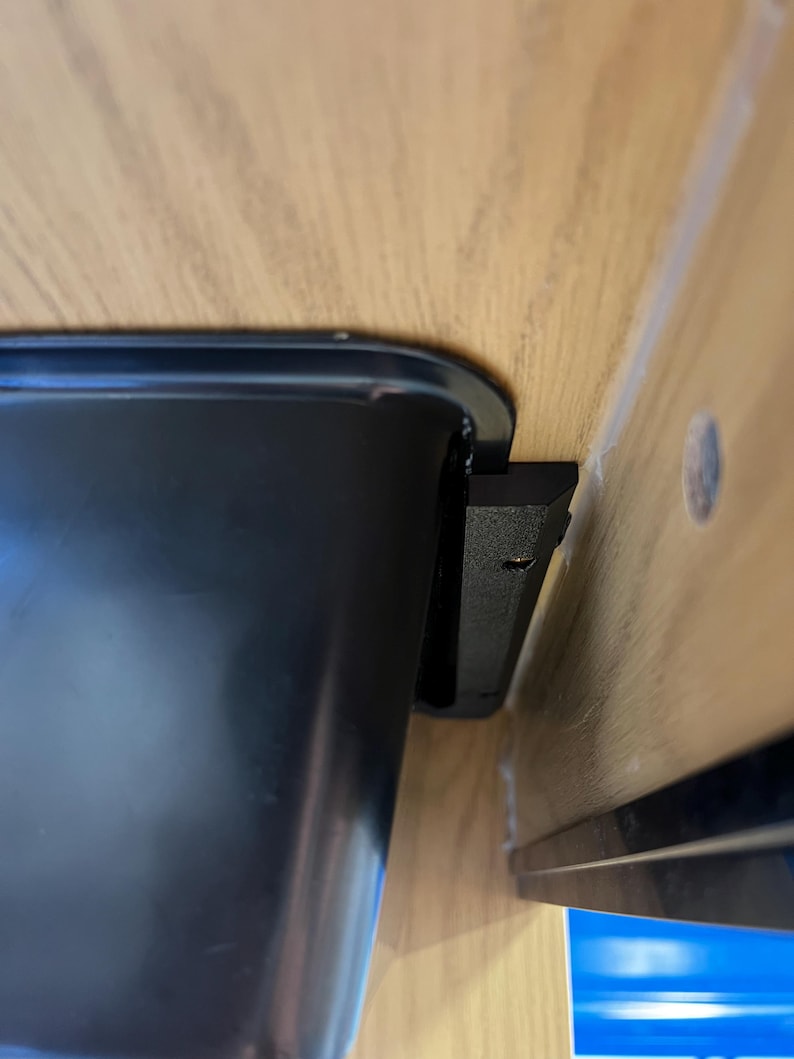 May include: A black, rectangular container with rounded corners is mounted under a wooden surface. A black bracket is attached to the container and the underside of the wood. The wood has a light brown color.