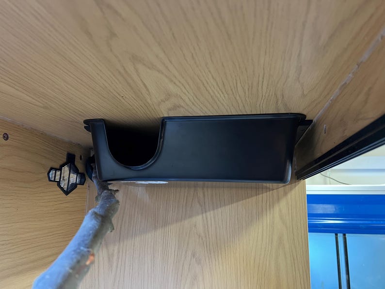 May include: A black, rectangular storage tray with a curved cutout, mounted under a light wood cabinet. The tray is designed to hold items and is attached to the cabinet with metal hardware. The cabinet has a wood grain pattern.